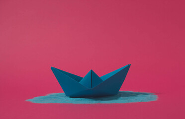 Blue origami paper boat floating on blue sand with pink background. Minimal concept.