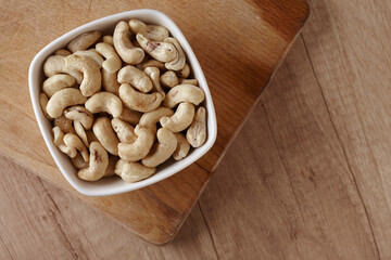 Tasty cashew nuts in bowl on a wooden cutting board on wooden table, top view. Space for text
