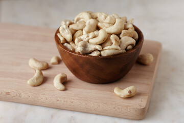 Tasty cashew nuts in bowl on a wooden cutting board on marble table, side view. Space for text