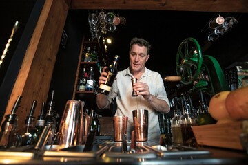 View of a professional bartender preparing a cocktail behind the counter of a beautiful and colourful cocktail bar using a shaker and bottles with fresh fruits in foreground, Lisbon, Portugal.