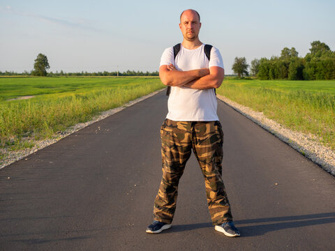 A Middle-aged Man With A Backpack On His Back Is Facing The Camera On The Road In The Field. His Hands Are Folded On His Chest