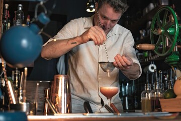 Professional bartender preparing a cocktail in a low glass using a shaker and a colander, view of a cheerful guy working in a fancy cocktail bar in night club in Lisbon, Portugal.