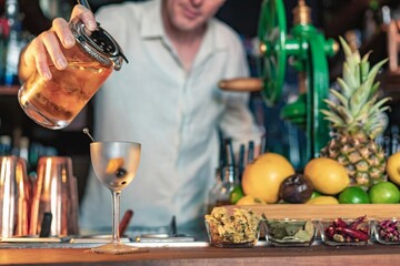 A professional bartender pouring a cocktail in a glass from a shaker and mixer, view of a barman working in a bar.