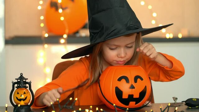Halloween. Happy little girl in witch costume and black hat fun brews potion from spiders, snakes, frogs in pumpkin pot.