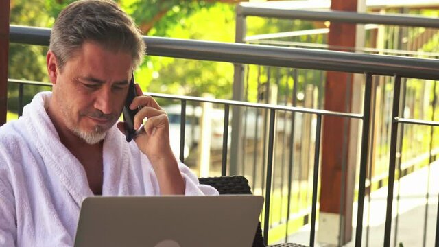 Man in bathrobe working at home in home office. Sitting on balcony outdoor using phone and laptop computer.