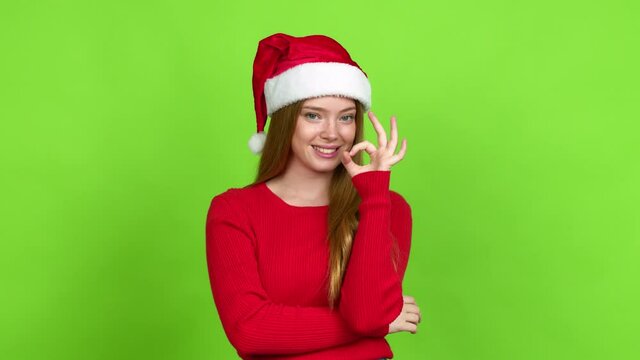 Teenager girl with christmas hat showing ok sign with fingers over isolated background. Green screen chroma key
