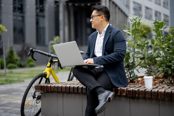 Asian businessman working on laptop computer on city street. Concept of modern successful man. Idea of remote and freelance work. Person wearing glasses and suit. Male sitting on bench