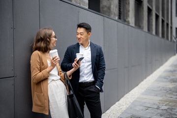 Asian businessman and caucasian businesswoman drinking coffee and talking in city. Concept of business cooperation. Idea of freelance and remote work. Woman with smartphone. Modern successful people