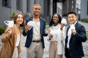 Multiracial business people holding medical masks and showing smartphone with app of Coronavirus Vaccination on city street. Concept of health protection during pandemic. Joyful young businessteam