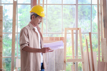 Elderly architect wearing helmet, holding blue print to inspects the structure, senior carpenter...