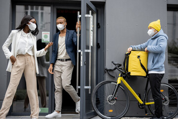 Courier with order waiting business people who coming out office building. Concept of shipping and logistics during Coronavirus pandemic. Idea of break on job. People wearing medical masks