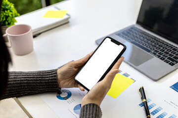 Businesswoman holding smartphone on desk with documents and laptop placed, she is looking at data from mobile phone, mobile phone blank screen for advertising graphics. The concept of using technology