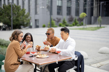 Multiracial business team having lunch with wine and pizza at outdoor cafe. Concept of teambuilding and corporate event. Idea of rest and leisure on job. People sitting at wooden table and talking