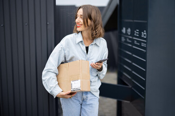 Young caucasian woman with parcel near automatic post terminal. Smiling girl holding smartphone and...