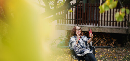 Fototapeta na wymiar Beautiful mature happy smiling woman chatting using smartphone online in autumn garden of her country house