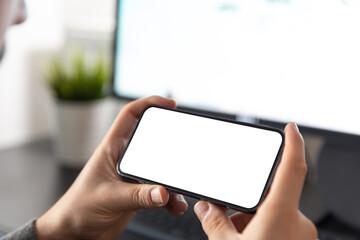 Man using smartphone blank screen frameless modern design while sitting in front of the computer in home office interior