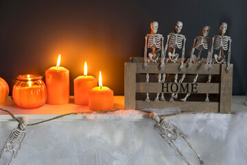Halloween home decoration. Plastic toy skeletons in a wooden box on a fireplace against a dark blue wall. A garland of skeletons. Cobweb on the dresser. Orange candles and lantern.