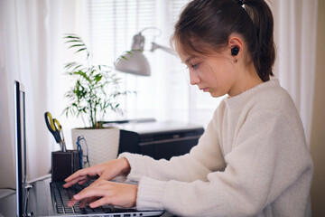 Young teenage girl working on a laptop, typing text at home in the workplace in a bright room.