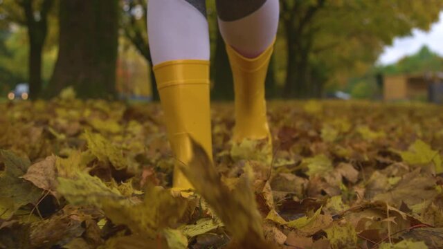 SLOW MOTION, LOW ANGLE, CLOSE UP, DOF: Young female wearing boots strolling around the park in autumn. Woman wearing yellow rubber boots kicks up dry fall colored leaves covering a grassy trail.