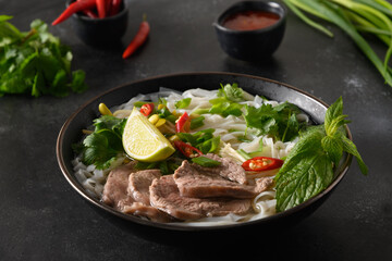 Pho Bo traditional Soup with beef, rice noodles, ginger, lime, chili pepper in bowl on black background. Close up. Vietnamese and Asian cuisine.