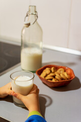 Child's hands grabbing a glass glass with milk next to a jar with cookies and a bottle on the kitchen counter.