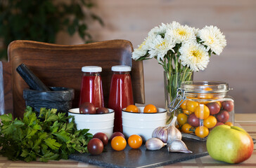 Still life. On the table are red and yellow cherry tomatoes, green parsley, garlic and apple, white asters. A wooden tray serves as a background. Tomato juice in two bottles. Canning.