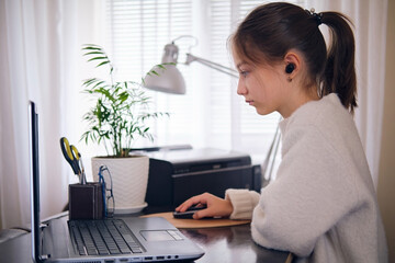 Young beautiful girl works at a laptop, holding a hand on a computer mouse at home at a work table in a bright room.