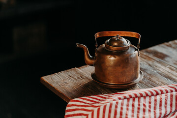 Horizontal shot of antique kettle on vintage wooden table. Old crockery. Metal copper teapot against dark background red towel near. Selective focus