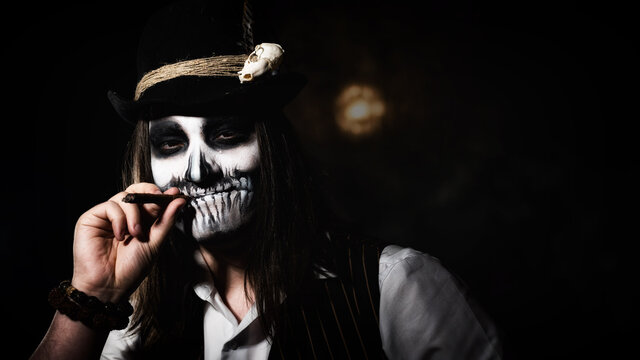 A young man in skull makeup smokes a cigar in the dark. Halloween skeleton costume
