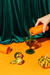 Orange pumpkins and a woman's hand pouring a beverage drink from an orange can against plush velvet curtain background. Creative Halloween and Thanksgiving concept. Contemporary fall still life idea.