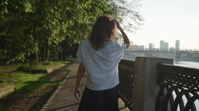 a brunette in casual clothes goes and touches hair in a city park near an openwork lattice. view from the back
