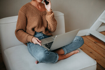 Young brunette girl with short hair in a brown sweater and blue jeans sitting on a white armchair and talking on a cell phone and holds a laptop in his hands