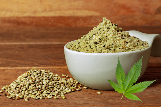 A pile of peeled hemp seeds in a white bowl with a hemp leafe and a pile of unpeeled henf seeds infront.