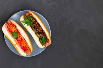 Plate with two hot dogs. Different tasty homemade hot dogs with tomato, cucumber, onion, salad, carrots, spices and mustard, ketchup on a dark table with copy space. flat lay. Fast food concept.