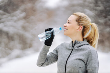 Happy sportswoman standing in nature at snowy winter day, taking a break and drinking fresh water....
