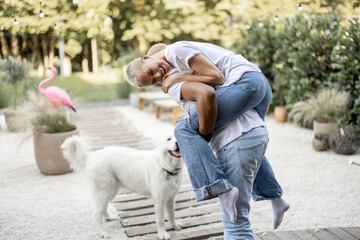 Latin boyfriend giving piggyback ride for his caucasian girlfriend on home terrace with a dog on the background. Concept of relationship and enjoying time together. Modern domestic lifestyle