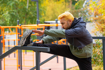 Man with dyed hair in blonde doing stretching on the sports ground outdoors. Open air training.