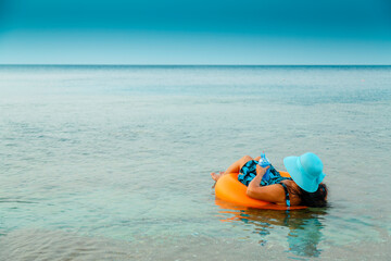 A woman in a straw hat and glasses in the sea in a swimming circle with a cocktail in her hand view from the back.