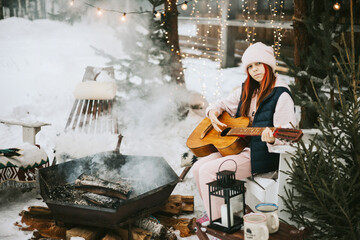 teenage girl in a vest and a knitted hat plays the guitar outdoor near a fire pit, the concept of...