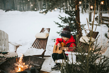 teenage girl in a vest and a knitted hat plays the guitar outdoor near a fire pit, the concept of...