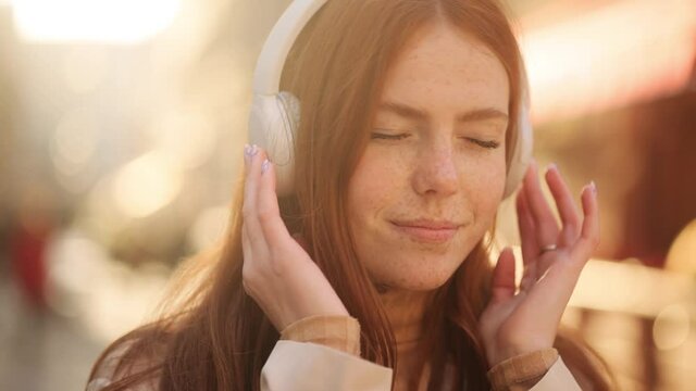 Close up of young millennial red head girl enjoying music and dancing in the city centre. Attractive young woman with freckles listening to music in headphones. High quality 4k footage