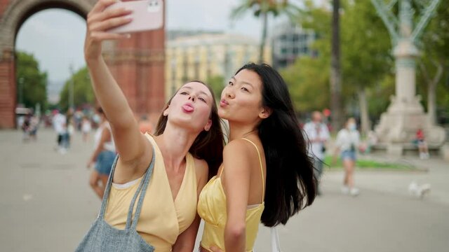 Two smiling diverse female friends looking at smartphone screen and making selfie portrait