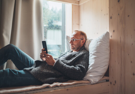 Smiling middle-aged man dressed warm knitted cardigan, jeans lying on cozy bed next to window and using modern smartphone. Everyday lifestyle photo with modern devices concept image.