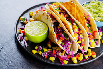 Vegetarian tacos with various vegetables, guacamole and lime on dark background. Tacos with sweet corn, purple cabbage and tomatoes on a black plate. Close-up