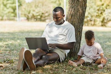 Fototapeta premium Full length portrait of young African-American man using laptop outdoors in park with cute baby son