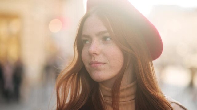 Sunny portrait of young beautiful caucasian red head woman in red beret looking at the camera at the city centre. Close up of happy young girl with freckles smiling. High quality 4k footage