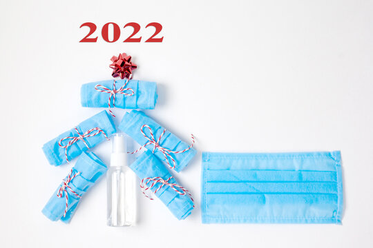 2022, Christmas and New year celebration concept. Photo of tree made of disposable mask, antiseptic decorated with red bow and number 2022 on white background. Flat lay with christmas gifts.