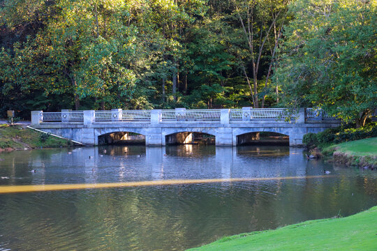 a stone bridge over the lake surrounded by green grass and lush green and autumn colored trees with still silky brown lake water at Lenox Park in Brookhaven Georgia USA