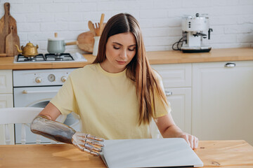Happy young woman with modern metal bionic arm works surfing internet with laptop sitting at wooden...