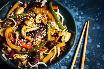 Wok with noodles, peppers, mushrooms, turkey meat and different vegetables and sesame seeds in a blue bowl on dark background